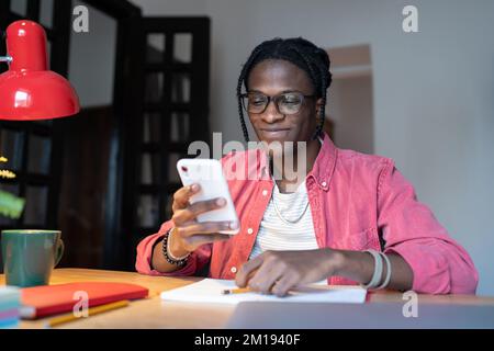 Ein glücklicher afrikanischer Student, der sich ein Smartphone ansieht und mit seiner Freundin chattet, während er zu Hause lernt Stockfoto