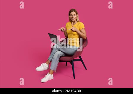 Fröhlich überrascht ziemlich reife, weiße, blonde Frau, die zwanglos auf einem Sessel sitzt und mit dem Finger auf den Laptop zeigt Stockfoto