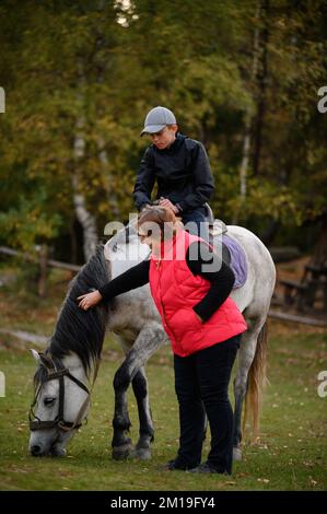 Großmutter und Enkel im Park, der Junge sitzt auf einem Pferd, und die Großmutter steht neben ihm, ein graues Pferd im Park. Stockfoto