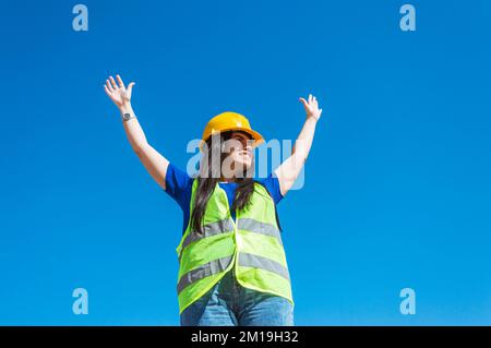 Eine weiße junge Frau, die als Ingenieurin verkleidet ist, trägt einen gelben Helm und blaue Kleidung. Sie steht mit offenen Armen auf der Straße Stockfoto