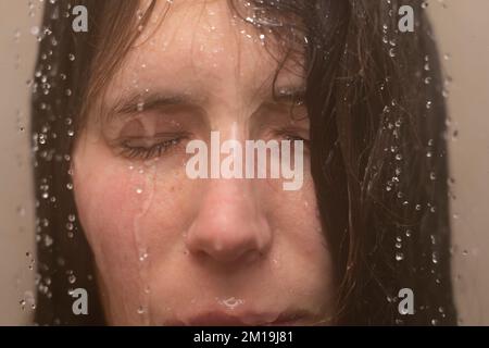 Eine junge Frau unter der Dusche, mit Wasser, das ihr ins Gesicht läuft. Konzept: Psychische Gesundheit, Ängste, Ängste, Gefühl gebrochen, körperdysmorphe Störung Stockfoto