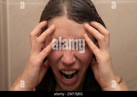 Eine junge Frau unter der Dusche, mit Wasser, das ihr emotionales Gesicht runterläuft. Konzept: Wütend, kann es nicht mehr ertragen, psychische Probleme, moderner Lebensstress Stockfoto