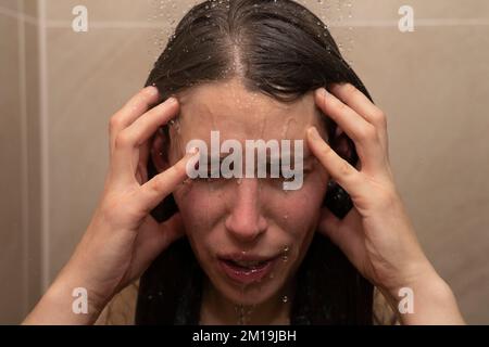 Junge Frau unter der Dusche, Wasser läuft ihr ins Gesicht und hält ihren Kopf. Konzept: Den Kopf halten, sich deprimiert fühlen, sich niedergeschlagen fühlen, Burn-out Stockfoto