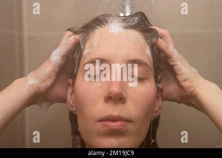 Eine Frau in den Dreißigern wäscht sich die Haare in der Dusche mit geschlossenen Augen und reibt sich Shampoo ins Haar Stockfoto