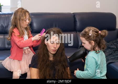 Eine Mutter und ihre beiden Töchter spielen und interagieren miteinander, wobei ein Mädchen die nassen Haare kämmt und das andere Kind die Haare mit einem Haartrockner trocknet Stockfoto