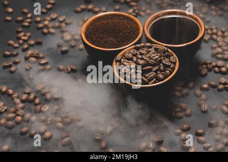 Tassen Kaffee unter ausgewählten und kalibrierten Arabica-Kaffeebohnen, verteilt auf schwarzem Hintergrund, drei aromatische Tassen Kaffee in einem Paar. Stockfoto
