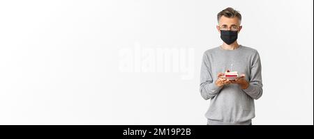 Konzept von Coronavirus, Quarantäne und Urlaub. Bild von traurigen Mann mittleren Alters in schwarzer medizinischer Maske und Brille, hält Geburtstagskuchen mit brennenden Kerze Stockfoto
