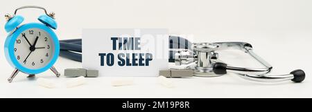 Medizin und Gesundheitskonzept. Auf einem isolierten weißen Hintergrund befinden sich eine Uhr, ein Stethoskop und eine Visitenkarte mit der Aufschrift "Zeit zum Schlafen" Stockfoto