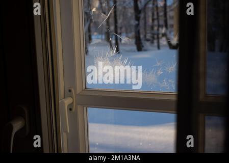 Eiszapfen auf einer Fensterscheibe an einem kalten Wintertag. Das Sonnenlicht scheint durch das Glas im Raum und erzeugt Schatten von Eisblumen auf dem Holz Stockfoto