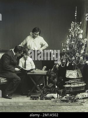 1915, Amsterdam, Niederlande. Altes Familienfoto einer Mutter und eines Vaters, das sich die Eröffnungsgeschenke ihres Sohnes neben einem weihnachtsbaum mit brennenden Kerzen ansieht Stockfoto