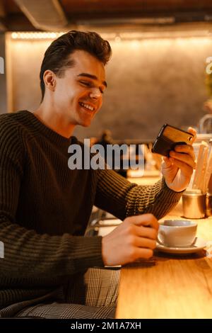 Ein junger, glücklicher weißer Mann, der einen Pullover trägt, Handy benutzt und Kaffee im Café trinkt Stockfoto