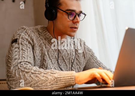 Junger weißer Mann mit Brille und lächelndem Lächeln, während er Laptop und Kopfhörer im Café benutzt Stockfoto