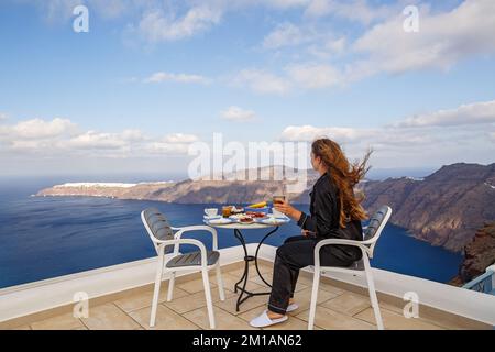 Die Frau sitzt und frühstückt an einem Tisch auf einer hohen Terrasse über dem Meer Stockfoto