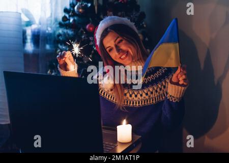 Feiern Sie Weihnachten und Neujahr online aus der Ferne mit einem Notebook. Eine Frau brennt zu Hause mit ukrainischer Flagge. Ich gratuliere der Familie Abroa Stockfoto