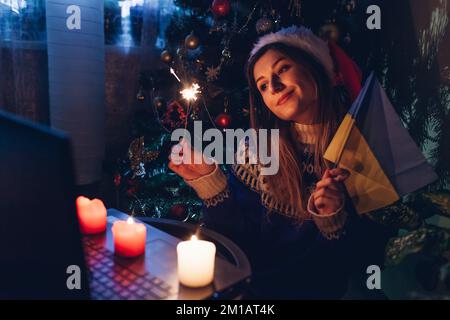 Die Frau verbrennt zu Hause an einem Baum mit ukrainischer Flagge. Feiern Sie Weihnachten und Neujahr in der Ukraine online aus der Ferne mit einem Laptop. Stockfoto