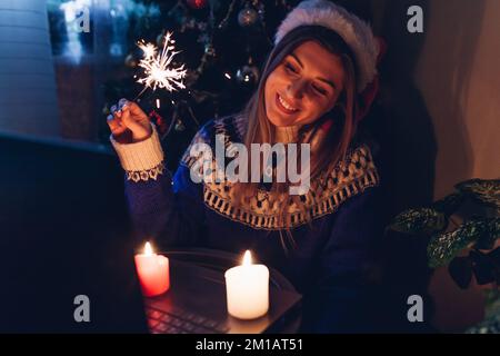 Feiern Sie Weihnachten und Neujahr online aus der Ferne mit einem Notebook. Eine junge Frau verbrennt zu Hause beim Telefonieren über das Internet an einem Weihnachtsbaum Stockfoto