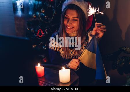 Wir feiern Weihnachten und Neujahr online aus der Ferne mit einem Laptop während des Krieges. Eine Frau brennt zu Hause mit ukrainischer Flagge. Ich rufe den Ehemann an Stockfoto