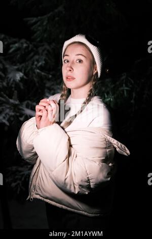 Eine junge, wunderschöne Blondine in Winterkleidung, die sich nachts in einem verschneiten Wald posiert, ein schöner frostiger Tag. Eine junge Frau wärmt ihre Hände Stockfoto