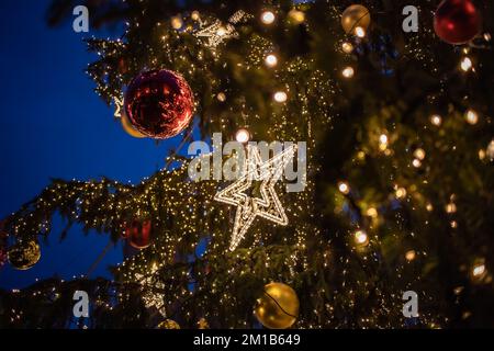 Nahaufnahme des äußeren Weihnachtsbaums mit festlichen Ornamenten am Abend. Nahaufnahme des dekorierten Weihnachtsbaums mit Kugel und Stern im Freien. Stockfoto