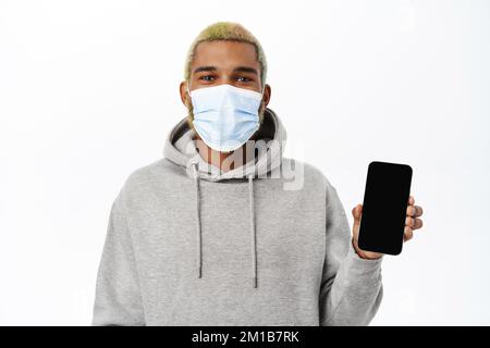 Covid 19, Coronavirus-Konzept. Lächelnder gutaussehender schwarzer Mann in medizinischer Gesichtsmaske, zeigt Smartphone-App, medizinische Handy-Anwendung Stockfoto