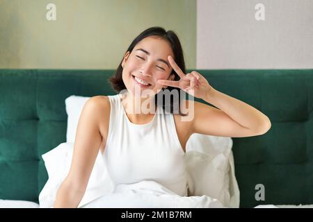 Positive asiatische Frau liegt im Bett, zeigt ein Friedenszeichen, genießt einen fröhlichen Morgen, wacht fröhlich auf, bleibt in ihrem Schlafzimmer Stockfoto