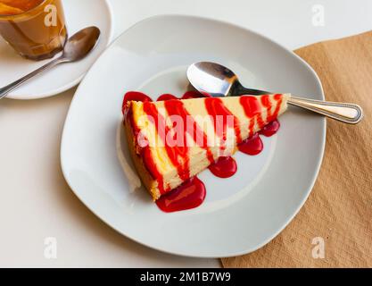 Ein Stück Käsekuchen mit frischer Erdbeermarmelade, serviert auf einem grauen Teller neben einem Glas Kaffee in der Cafeteria Stockfoto