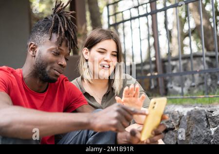 Multirassisches junges Paar, das draußen mit Mobiltelefonen sitzt - ein amerikanisches, nettes junges Paar, das Videos auf dem Handy ansieht Stockfoto
