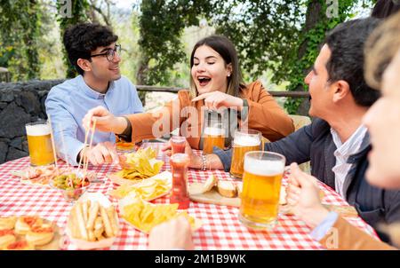 Eine Gruppe von gemischten Freunden, die Bier trinken und Tapas essen - glückliche Leute, die zusammen Spaß im Garten des Bauernhofs haben Stockfoto