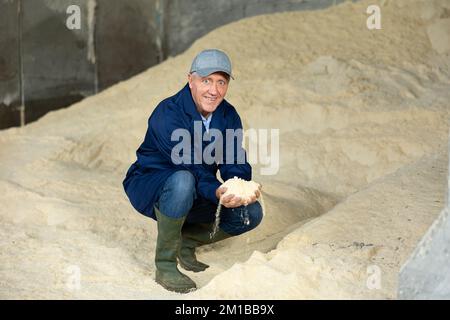 Positiver Bauer, der auf einem Haufen Maismehl und Tierfutter hockt Stockfoto