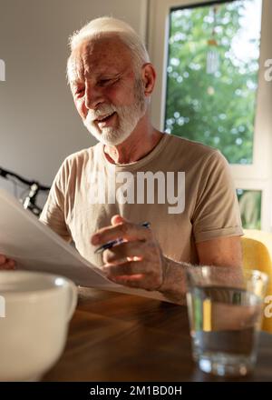 Zufriedener Seniorenmann, der zu Hause am Tisch sitzt und mit einem Lächeln im Gesicht Papier liest Stockfoto