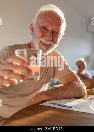 Glücklicher Senior-Mann, der zu Hause am Esstisch sitzt und ein Glas Wasser und Prost. hält Gesunder Lebensstil im Alter Stockfoto