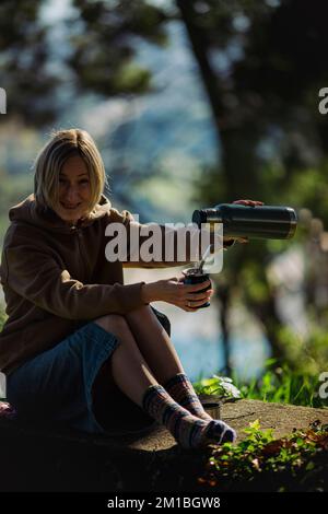 Eine Frau im Park, die heißes Wasser aus einer Thermoskanne in einen Mate Becher gießt. Stockfoto