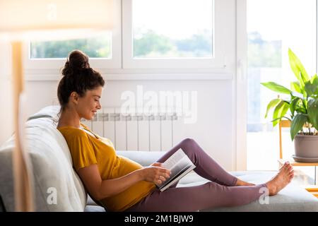 Hübsche schwangere Frau, die auf dem Sofa sitzt, sich ausruht und ein Buch auf dem Sofa im Wohnzimmer liest Stockfoto