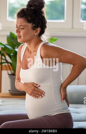 Schwangere Frau, die auf dem Sofa sitzt und Rücken und Bauch mit Schmerzausdruck im Gesicht hält Stockfoto