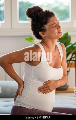 Schwangere Frau, die auf dem Sofa sitzt und Rücken und Bauch mit Schmerzausdruck im Gesicht hält Stockfoto
