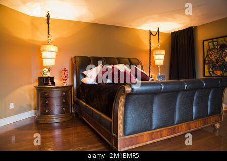 Kingsize-Bett aus Holz und Leder, Schlittenbett und Nachttisch im Hauptschlafzimmer mit braunen gelb bemalten Wänden im oberen Stockwerk im luxuriösen Haus. Stockfoto