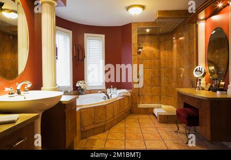 Weißes Sockelwaschbecken und Schminktisch mit roter Samtbank und Terrakotta-Keramikbadewanne im geräumigen Hauptbadezimmer im luxuriösen Haus. Stockfoto