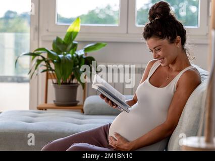 Hübsche schwangere Frau, die auf dem Sofa sitzt, sich ausruht und ein Buch auf dem Sofa im Wohnzimmer liest Stockfoto