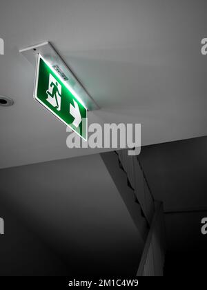 Notausgangsschild. Grünes Notausgangsschild, das an der weißen Decke hängt und auf dem dunklen Flur in der Nähe der Treppenleitertür im Gebäude leuchtet, Stockfoto
