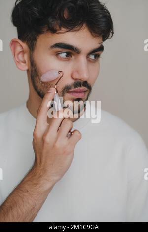 Zugeschnittenes Bild eines Mannes, der sein Gesicht mit Jadewalze massiert. Körper- und Hautpflege. Isoliert auf weißem Hintergrund. Studioaufnahmen Stockfoto