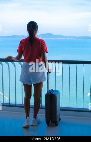 Asiatische Frauen mit Handgepäck und einem Trolley, die in einem Hotelzimmer mit Blick auf das Meer in Thailand einchecken. Stockfoto