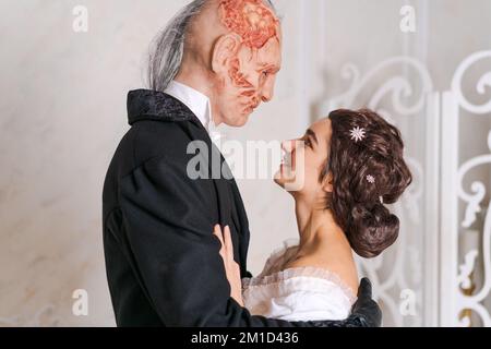 Hübsches Mädchen in weißem Kleid und gruseliger entstellter Mann in schwarzem Stand in Umarmung. Geister-Cosplay, halloween-Feiertagskonzept Stockfoto