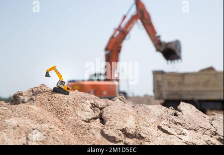 Konzentriere dich auf einen kleinen gelben Bagger, der auf dem Boden steht. Im Hintergrund, unscharf, große Arbeitsmaschinen - ein Bagger und ein Lkw bei einem Stockfoto
