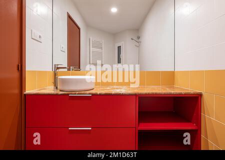 Weißes Spüle über Kopf in einem roten Schrank in einem hellen weißen und gelben Badezimmer Weiße und gelbe Fliesen an der Wand und brauner Boden aus Porzellan-Steingut Stockfoto