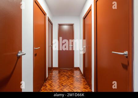 Hell erleuchteter langer Flur mit vielen Türen in derselben dunkelorangefarbenen Farbe, die zu separaten Apartments führen. Der Boden ist Parkett mit abstraktem Muster Stockfoto
