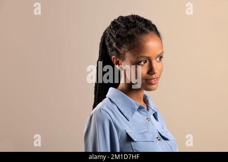 Junges schwarzes weibliches Modell mit Afro-Geflechten und blauem Hemd, das auf beigefarbenem Hintergrund steht und wegblickt Stockfoto