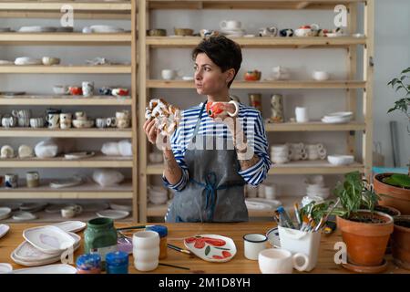 Hungrige Töpferkünstlerin in der Schürze frühstückt, während sie allein in ihrem Keramikladen steht Stockfoto