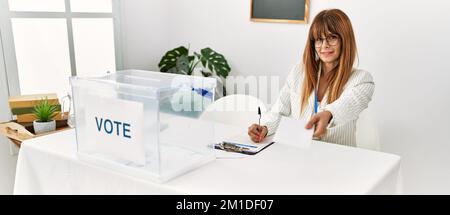 Junge hispanische Frau lächelt selbstbewusst und hält Stimmabgabe und schreibt in die Zwischenablage am Wahlkollegium Stockfoto