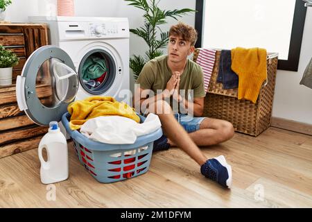 Junger weißer Mann, der schmutzige Wäsche in die Waschmaschine legt, bettelt und betet mit Händen zusammen, mit Hoffnung Ausdruck im Gesicht sehr emotional und Stockfoto