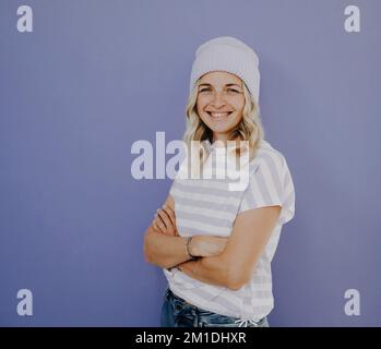Schöne Frau mit Mütze und gereiftem Hemd steht vor lila Hintergrund Stockfoto